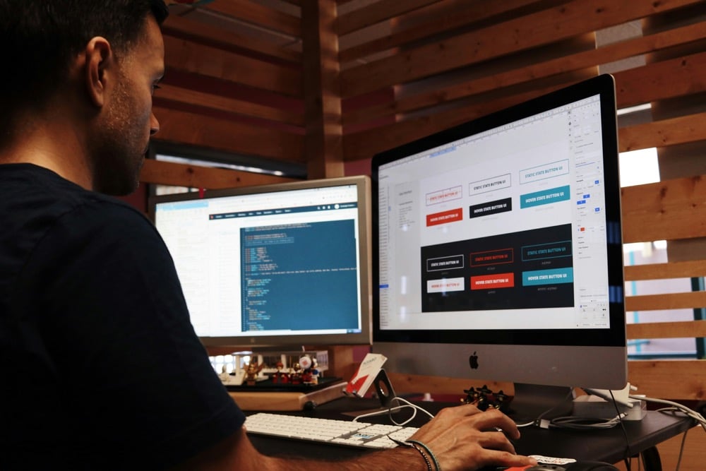 A person works at a desk with two monitors, one displaying code and the other showing a user interface design with colored buttons.