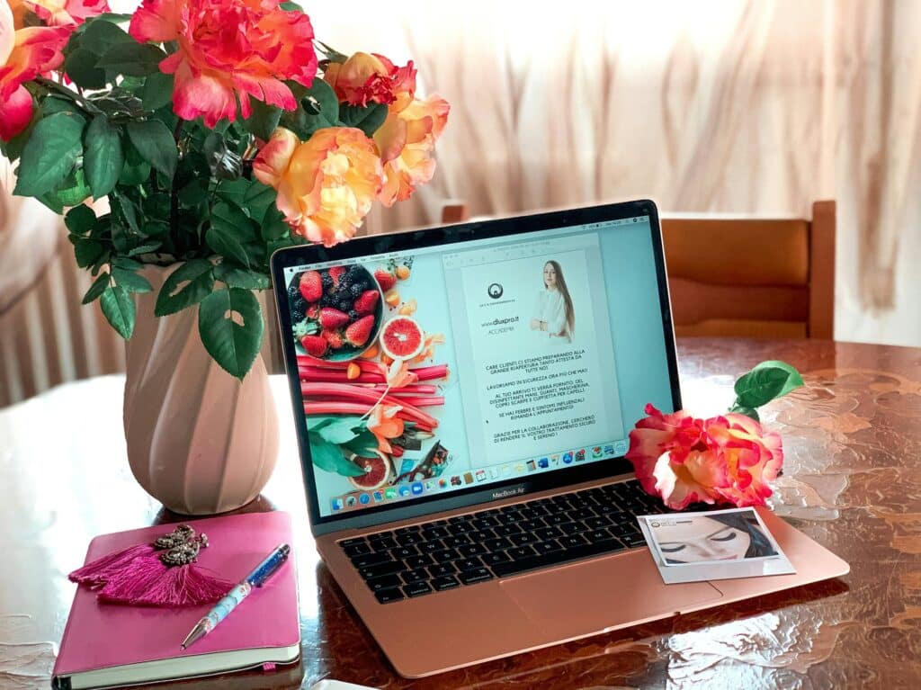 A laptop displaying a digital magazine sits on a table with a vase of flowers, a pink notebook, a pen, a photo print, and a flower next to it.